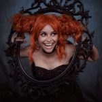 A lively portrait of a woman with orange hair posing through a decorative black frame, captured indoors. — Chatroulette O
